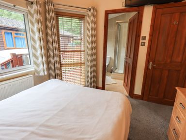 A bedroom with a bed covered with white bedding two windows with curtains and blinds a door leading to a bathroom with a shower and toilet two wooden doors and a dresser at Thirlmere View Thirlmere 6 White Cross Bay near Windermere