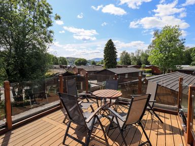 An outdoor wooden deck with a round table and six mesh chairs overlooking wooden cabins and trees at Thirlmere View Thirlmere 6 White Cross Bay near Windermere