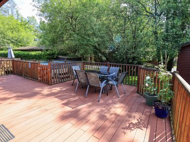 An outdoor wooden deck with a glass table and six chairs surrounded by wooden railings and green trees at Thirlmere View Thirlmere 6 White Cross Bay near Windermere
