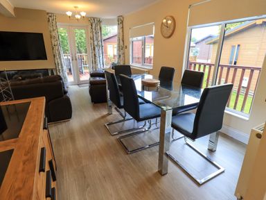 A dining area with a glass table and six black chairs next to a sitting area with brown sofas and a TV at Thirlmere View in Thirlmere 6 White Cross Bay near Windermere