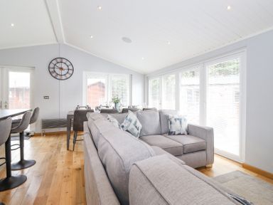 A living area with grey sofas, bar stools, a dining table, and large windows at Lakes Lodge Keswick Court 3 White Cross Bay near Windermere