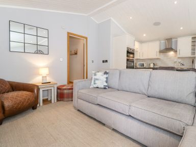 A living room with a grey sofa a leather armchair a side table with a lamp and a kitchen area at Lakes Lodge Keswick Court 3 White Cross Bay near Windermere