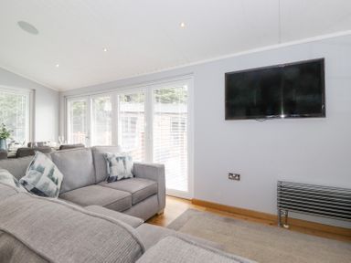 A living room with a grey sofa a wall-mounted television and large windows with blinds at Lakes Lodge Keswick Court 3 White Cross Bay near Windermere