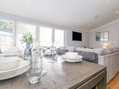 A dining table set with white dishes and glasses next to a living area with gray sofas and a wall-mounted TV at Lakes Lodge in Keswick Court 3 White Cross Bay near Windermere