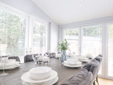 A dining table set with white plates bowls and glasses in a room with large windows at Lakes Lodge in Keswick Court 3 White Cross Bay near Windermere