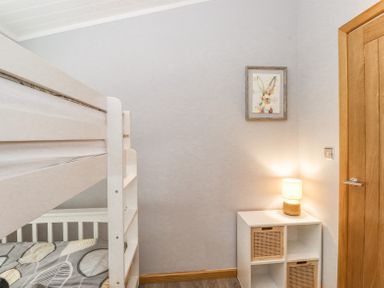 A bedroom with a white bunk bed a small white storage unit with wicker drawers a lamp on top and a framed picture of a rabbit on the wall at Lakes Lodge Keswick Court 3 White Cross Bay near Windermere