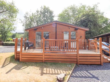A wooden cabin with a deck and outdoor chairs surrounded by trees at Lakes Lodge Keswick Court 3 White Cross Bay near Windermere
