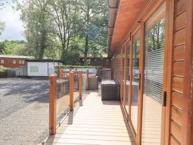 An outdoor wooden deck with glass and wood railing adjacent to a wooden building with glass doors and outdoor furniture at Lakes Lodge in Keswick Court 3 White Cross Bay near Windermere