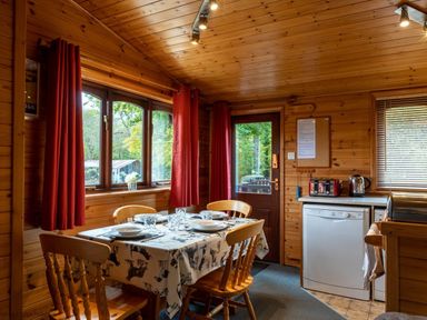 A wooden dining room with a table set for four next to windows with red curtains and a door near kitchen appliances at Glaramara Number 7 Burnside Park near Keswick