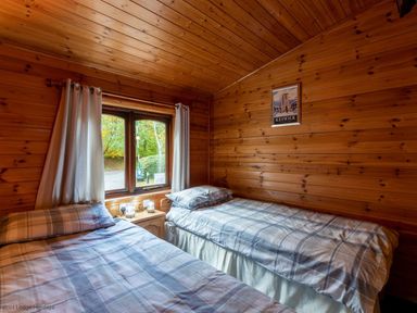 A bedroom with two single beds a window with curtains a small wooden nightstand with lamps and a framed picture on the wooden wall at Glaramara Number 7 Burnside Park near Keswick