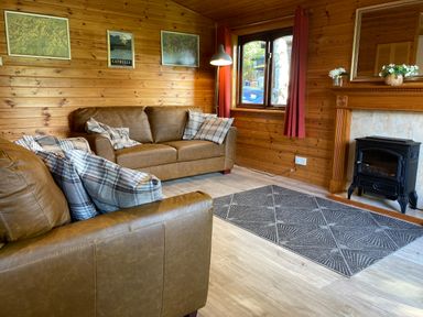 A living room with brown leather sofas and plaid cushions wood panel walls a window with red curtains a fireplace with flowers and a black stove at Glaramara Number 7 Burnside Park near Keswick