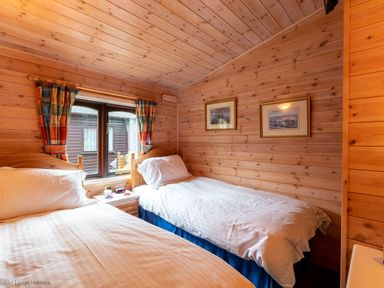 A bedroom with two single beds a window with colorful curtains and two framed pictures on wooden walls at Rivendell Burnside Park Number 14 Burnside Park near Keswick