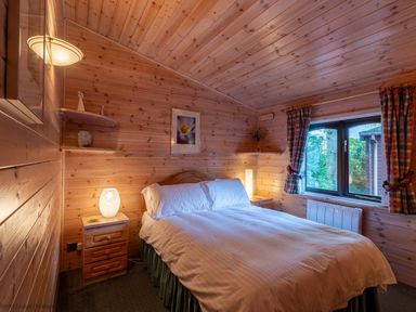 A bedroom with a double bed wooden walls a window with curtains and bedside tables with lamps at Rivendell Burnside Park Number 14 Burnside Park near Keswick