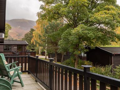 A wooden balcony with green chairs overlooking trees and dark wooden lodges with a mountain in the background at Rivendell Burnside Park Number 14 Burnside Park near Keswick