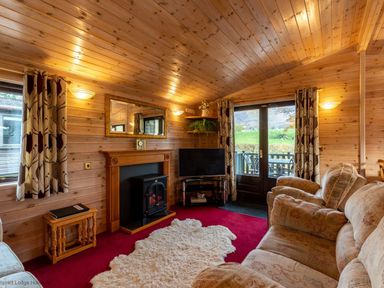 A living room with wooden walls and ceiling a fireplace a television sofas and sliding doors leading outside at Burr Ghyll Number 27 Burnside Park near Keswick