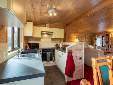 A kitchen with sink oven microwave and red towels next to a living room with sofas and window at Burr Ghyll Number 27 Burnside Park near Keswick
