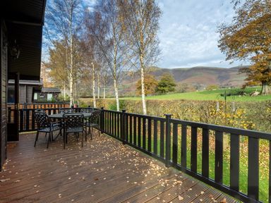 A wooden deck with a black metal table and six chairs overlooking trees and hills at Burr Ghyll Number 27 Burnside Park near Keswick