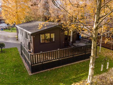 A wooden cabin with a fenced porch and patio furniture surrounded by autumn trees at Burr Ghyll Number 27 Burnside Park near Keswick