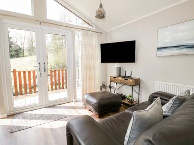 A living room with leather sofa and ottoman by glass doors and TV on wall at Troutbeck Lodge in Beckside 6 Limefitt Park near Windermere