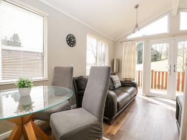 A living area with a glass round table and two chairs a black leather sofa and large windows with blinds at Troutbeck Lodge Beckside 6 Limefitt Park near Windermere