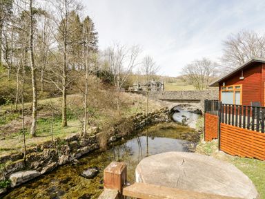 A wooden cabin beside a stream with a stone bridge and leafless trees at Troutbeck Lodge in Beckside 6 Limefitt Park near Windermere