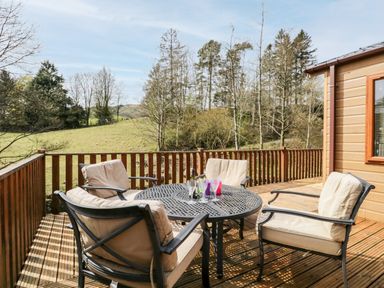 An outdoor wooden deck with a metal round table and four cushioned chairs overlooking a grassy field and trees at Troutbeck Lodge in Beckside 6 Limefitt Park near Windermere