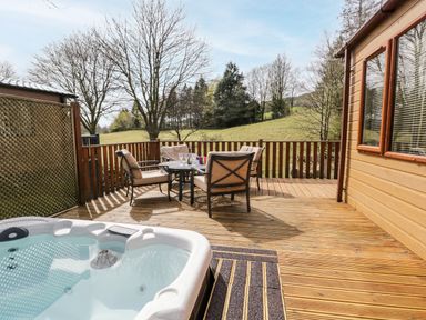 An outdoor wooden deck with a hot tub and a table with four chairs and a rural landscape at Troutbeck Lodge Beckside 6 Limefitt Park near Windermere