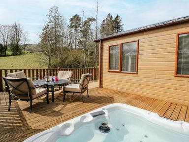 An outdoor wooden deck with a round table and four cushioned chairs next to a hot tub and a wooden cabin Beckside 6 Limefitt Park near Windermere