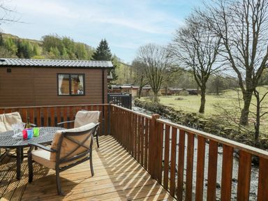 A wooden deck with a round table and cushioned chairs next to a cabin and overlooking a grassy area with trees and a stream at Troutbeck Lodge Beckside 6 Limefitt Park near Windermere