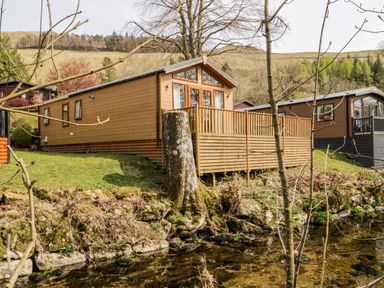 A wooden lodge with a deck near a stream in a rural area at Troutbeck Lodge Beckside 6 Limefitt Park near Windermere