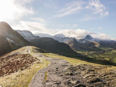 A mountain trail with rocky and grassy terrain overlooking snow-capped peaks and green valleys in clear weather