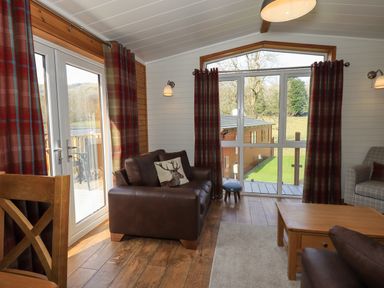 A living room with a leather sofa a wooden table plaid curtains and large windows at Ramblers Rest Lodge Pinewood 22 Limefitt Park near Windermere