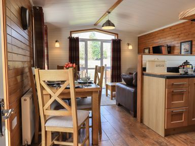 A dining area with wooden chairs and table set with plates and glasses next to a living area with a brown leather sofa and kitchen counter at Ramblers' Rest Lodge Pinewood 22 Limefitt Park near Windermere