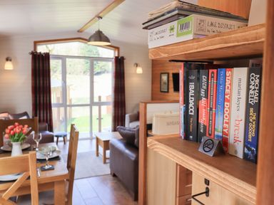 A close-up of a bookshelf with books and a jigsaw puzzle next to a dining area and living room with a table set and curtain-covered windows at Ramblers Rest Lodge Pinewood 22 Limefitt Park near Windermere