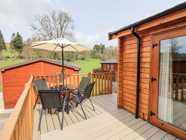 An outdoor deck with a round table four chairs and an umbrella next to wooden cabins at Ramblers Rest Lodge Pinewood 22 Limefitt Park near Windermere