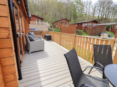 A wooden deck with chairs and table outside wooden lodges surrounded by trees at Ramblers Rest Lodge Pinewood 22 Limefitt Park near Windermere