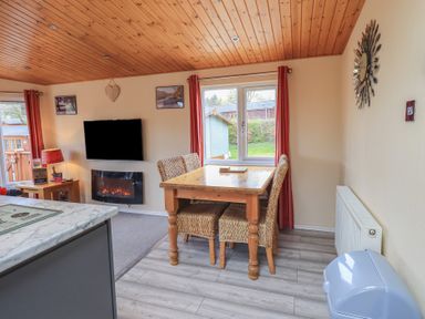 A dining area with a wooden table and wicker chairs near a window and a living space with a wall-mounted TV and electric fireplace at Striding Edge Lodge Ullswater 9 Limefitt Park near Windermere