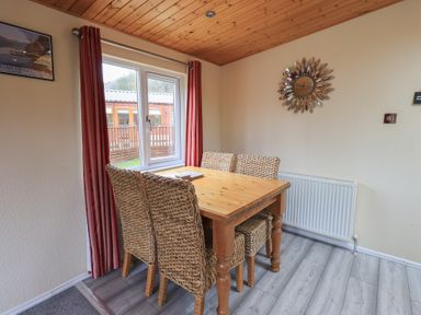 A dining area with a wooden table and four wicker chairs near a window with red curtains at Striding Edge Lodge Ullswater 9 Limefitt Park near Windermere