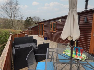 An outdoor wooden deck with a black wicker sofa set and a metal table with blue chairs at Striding Edge Lodge in Ullswater 9 Limefitt Park near Windermere