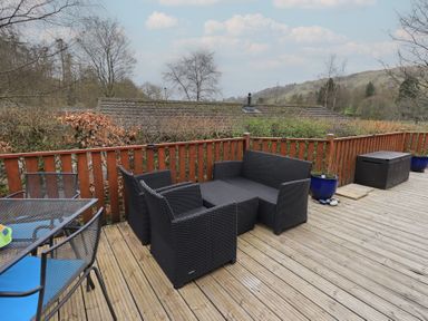 An outdoor wooden deck with black wicker seating a glass table with blue chairs blue plant pots and a wooden storage box at Striding Edge Lodge Ullswater 9 Limefitt Park near Windermere