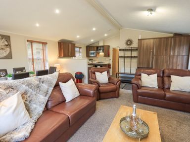 A living room with brown leather sofas and armchair a coffee table with a bottle and glasses and an open kitchen with wooden cabinets at Horseshoe Lodge Wansfell 5 Limefitt Park near Windermere