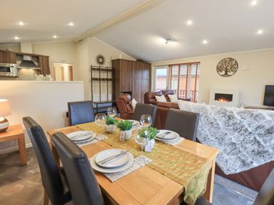 A dining table set with plates, cutlery, glasses and plants next to a living area with brown leather sofas and a fireplace at Horseshoe Lodge in Wansfell 5, Limefitt Park near Windermere