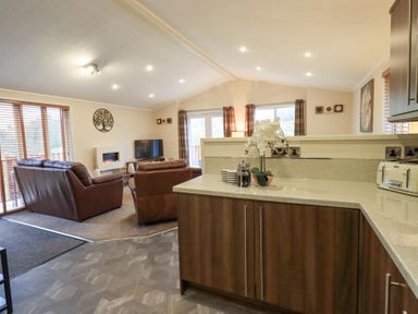 A living room with brown leather sofas a television a fireplace and wooden blinds viewed from a kitchen area with a countertop and cabinets at Horseshoe Lodge Wansfell 5 Limefitt Park near Windermere