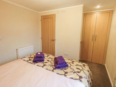 A bedroom with a bed with patterned bedding and two sets of folded purple towels on it a wooden door a radiator and a built-in wooden wardrobe at Horseshoe Lodge in Wansfell 5 Limefitt Park near Windermere