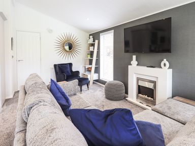 A living room with a gray sofa and blue cushions a black armchair a fireplace a wall-mounted television and a decorative mirror at Jinnyspinner Lodge Beckside 37 Limefitt Park near Windermere