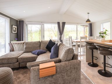 A living room with grey sectional sofa and blue cushions near windows with curtains and a dining area with white chairs and a kitchen bar with stools at Jinnyspinner Lodge Beckside 37 Limefitt Park near Windermere