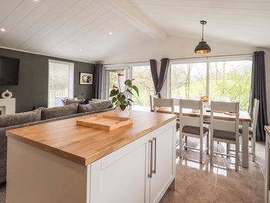 A dining area with a wooden table and chairs next to large windows and a kitchen island with a wooden countertop and a plant on it at Jinnyspinner Lodge Beckside 37 Limefitt Park near Windermere