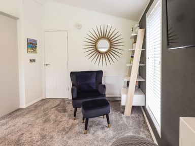 A room with a black armchair and matching footstool on grey carpet with a sunburst mirror on the wall at Jinnyspinner Lodge Beckside 37 Limefitt Park near Windermere
