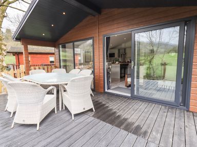 An outdoor wooden deck with a glass table and white wicker chairs outside a cabin with sliding glass doors at Jinnyspinner Lodge Beckside 37 Limefitt Park near Windermere