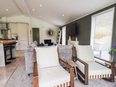A living area with two wooden chairs with white cushions a grey sofa a wall-mounted television and a kitchen area with bar stools at Jinnyspinner Lodge Beckside 37 Limefitt Park near Windermere